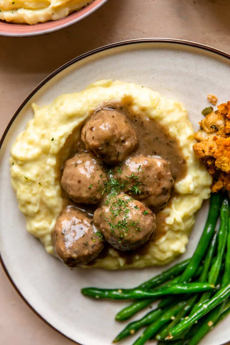 Turkey meatballs and gravy served over mashed potatoes on a plate with green beans and stuffing on the side.
