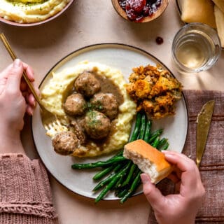 Hands around a plate of turkey meatballs served over mashed potatoes, bread rolls, green beans and stuffing.