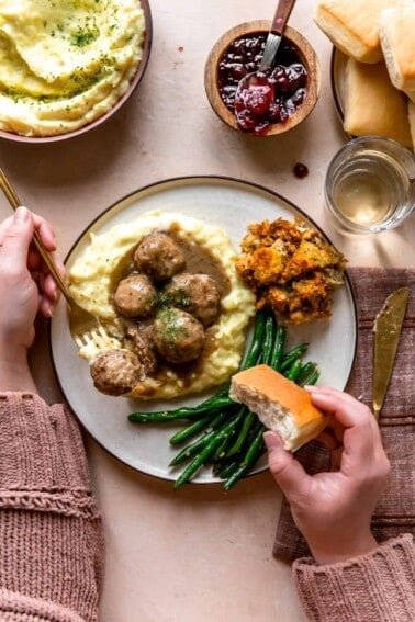 Hands around a plate of turkey meatballs served over mashed potatoes, bread rolls, green beans and stuffing.