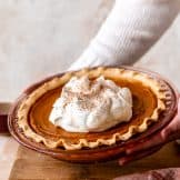 Hands holding up a sweet potato pie topped with fluffy whipped cream and sprinkled with ground nutmeg.