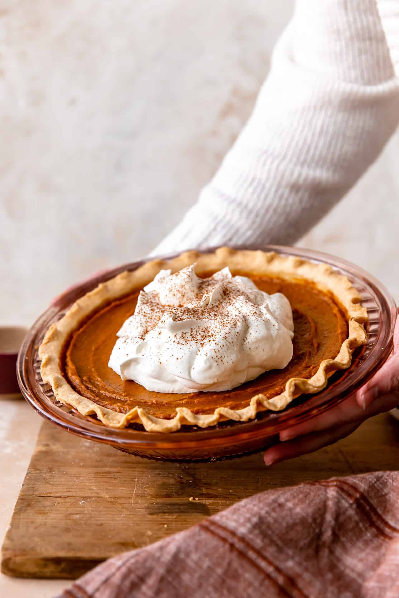 Hands holding up a sweet potato pie topped with fluffy whipped cream and sprinkled with ground nutmeg. 