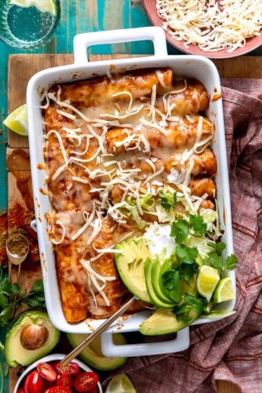 White baking dish filled with chicken enchiladas and topped with sliced avocado, sour cream and fresh cilantro.