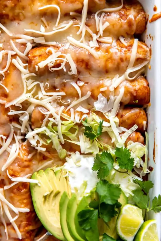 Up close view of enchiladas topped with melted cheese, sliced avocado, sour cream and lime wedges.
