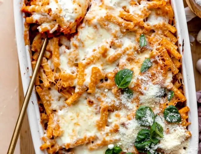 Spoon dishing into a baking dish filled with baked ziti, topped with melted cheese and basil leaves.
