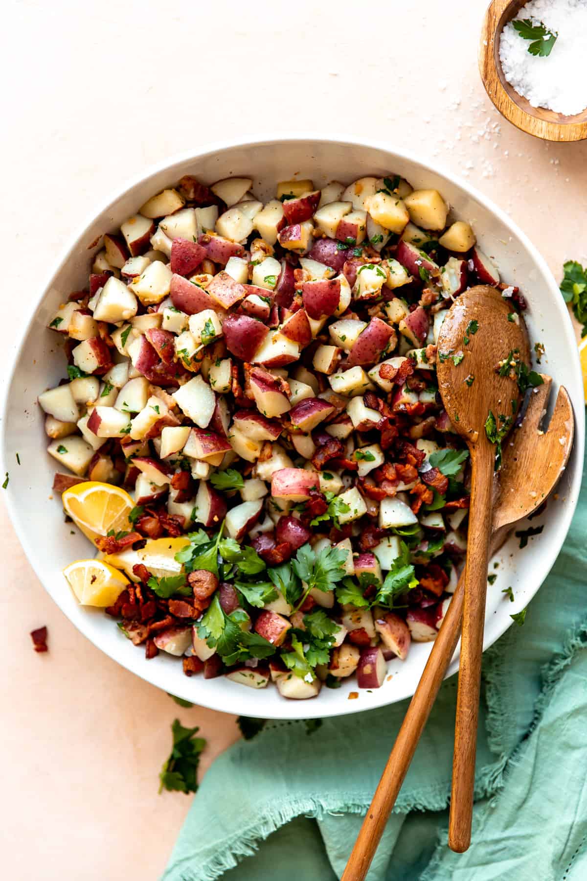 White bowl on a teal napkin, serving warm german potato salad with wooden spoons.