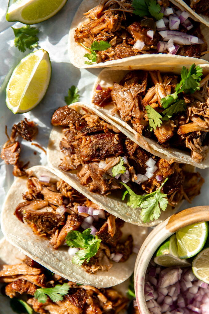 Carnitas Tacos lined up and topped with fresh cilantro, and diced red onion, lime wedges on the side.