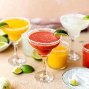 Frozen fruit margaritas lined up with mango, strawberry, and lime, with wedges of limes surrounding.