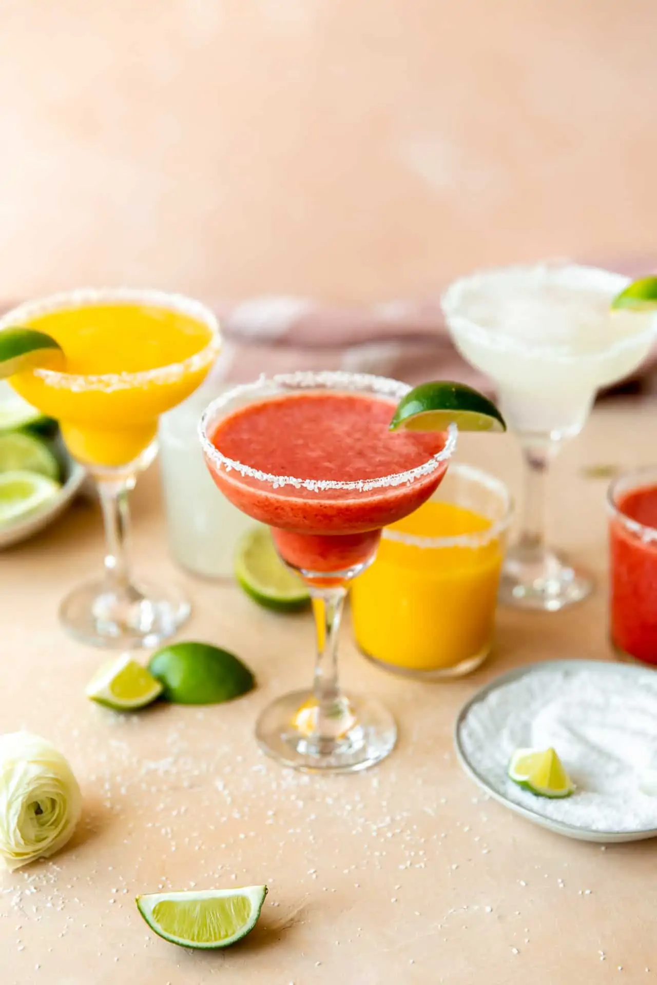 Frozen fruit margaritas lined up with mango, strawberry, and lime, with wedges of limes surrounding.