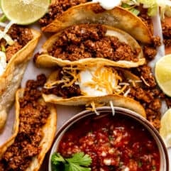 Plate of ground beef tacos on fried corn tortillas topped with sour cream and served with salsa.