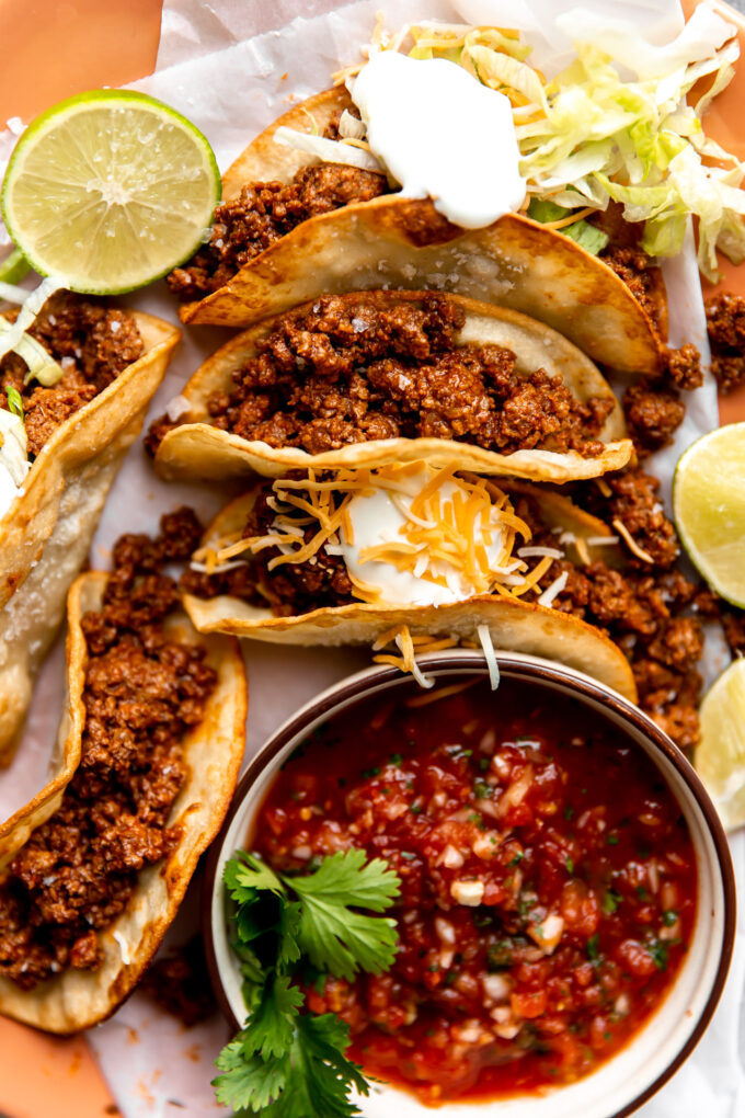 Plate of ground beef tacos on fried corn tortillas topped with sour cream and served with salsa.