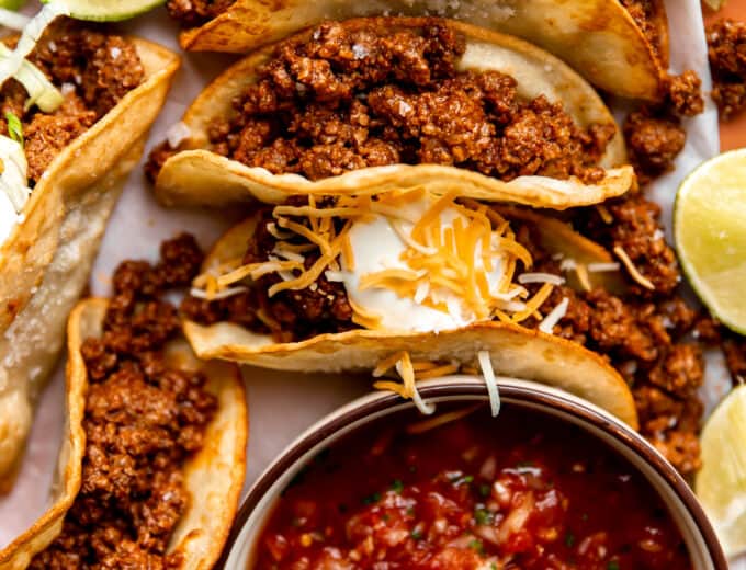 Plate of ground beef tacos on fried corn tortillas topped with sour cream and served with salsa.