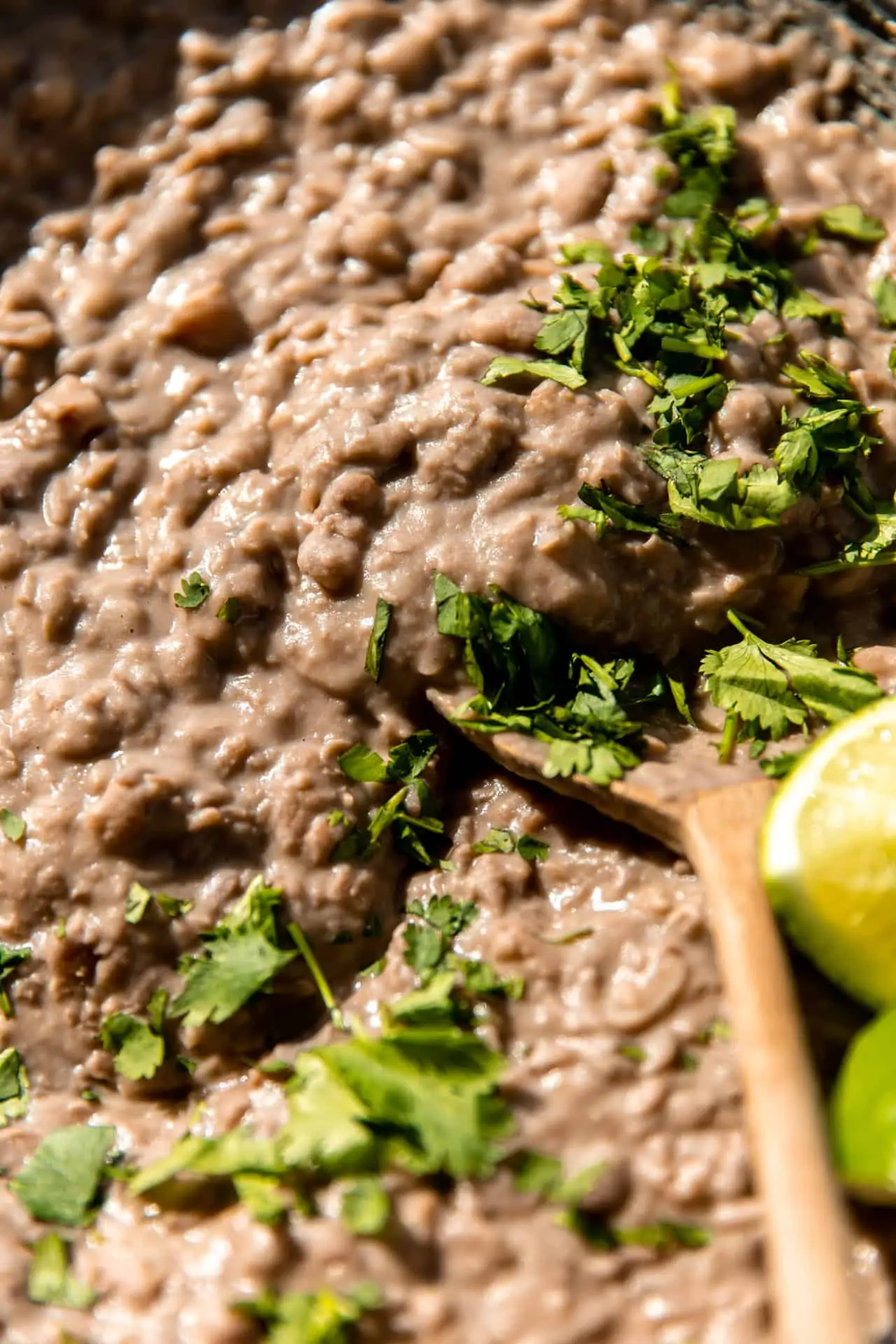 Spoon scooping up thick and creamy refried beans topped with cilantro and lime wedges.