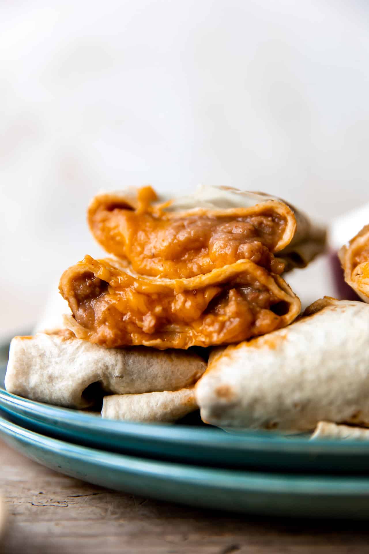 Bean and cheese burritos stacked on a plate showing melty cheese and creamy beans with red sauce inside.