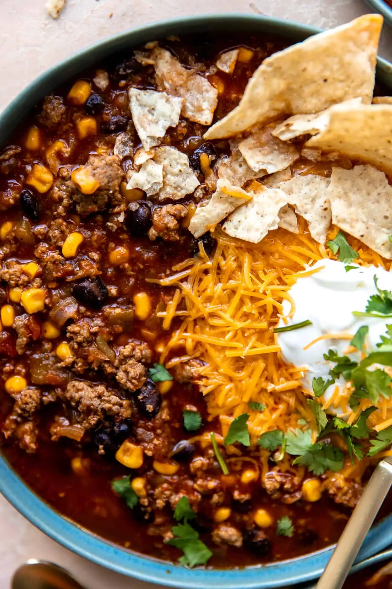 Up close view of a bowl of taco soup showing chunks of beef, beans and corn in a dark red broth topped with melted cheese and sour cream.