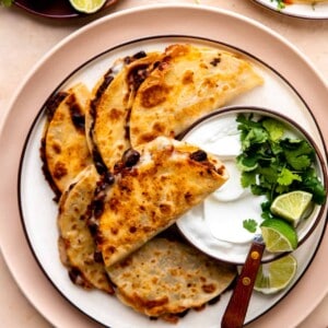 Plate filled with crispy black bean tacos served with a bowl of sour cream, cilantro and lime.