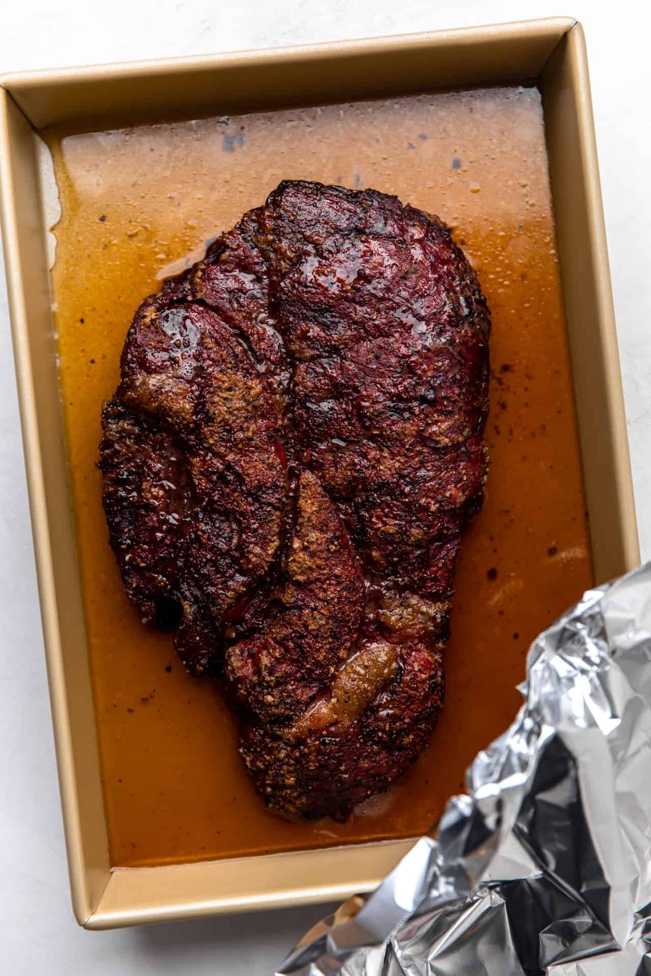 Smoked chuck roast in a baking tray with a small amount of broth and foil for braising.