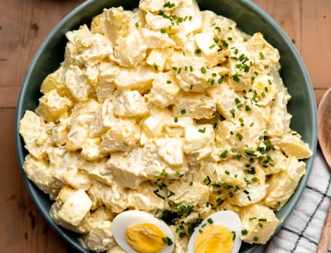 Bowl of creamy potato salad made with egg and dill pickle. Topped with sliced apart hard boiled egg.