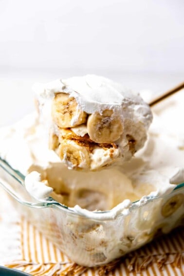 Banana pudding in a baking dish being scooped up showing sliced bananas and nilla wafers with pudding and whipped cream.