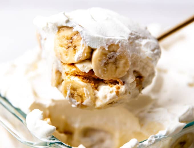 Banana pudding in a baking dish being scooped up showing sliced bananas and nilla wafers with pudding and whipped cream.