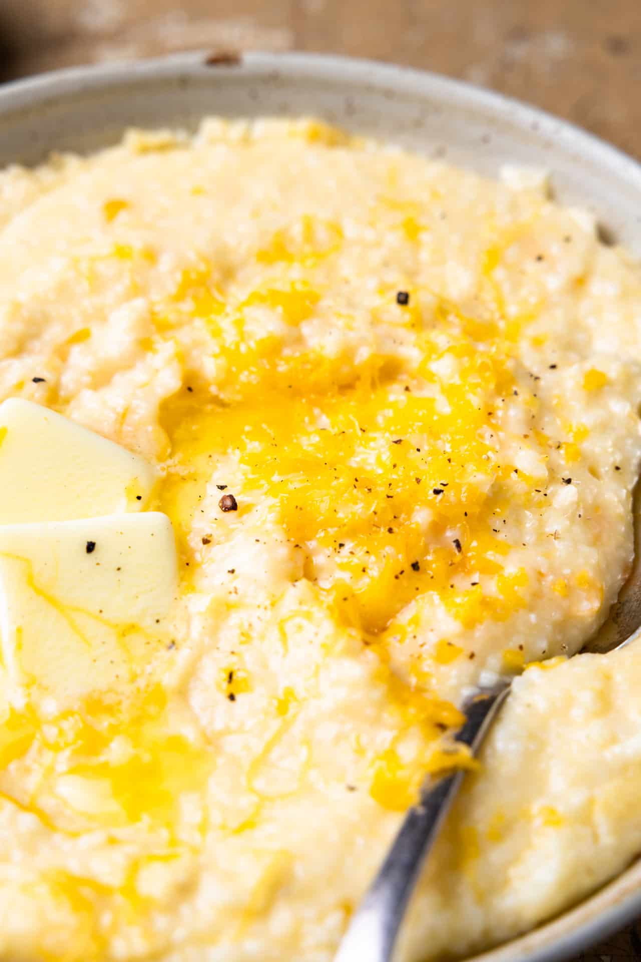 Up close showing texture of cooked cheese grits, topped with butter and fresh pepper. 