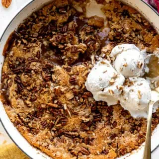 baking dish with pecan cobbler showing caramel sauce and topped with ice cream.