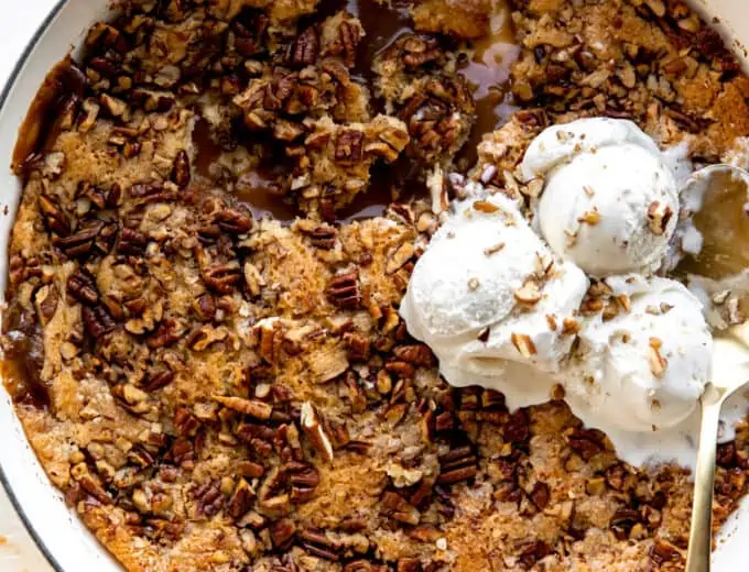 baking dish with pecan cobbler showing caramel sauce and topped with ice cream.