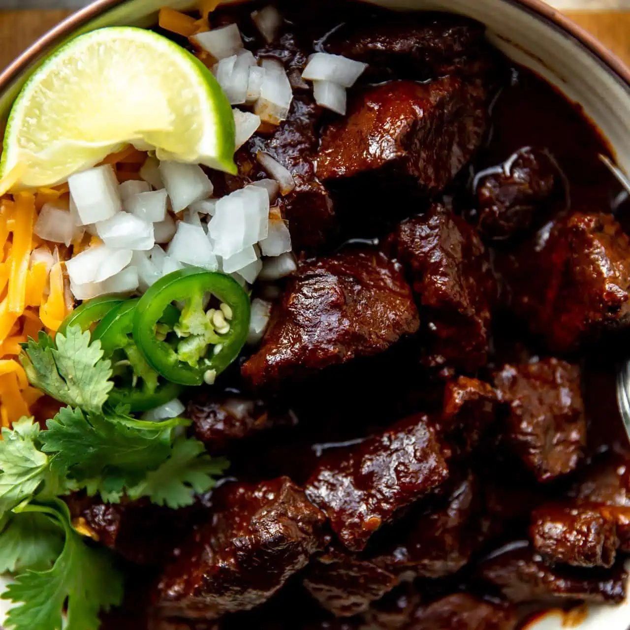 Bowl filled with Authentic Texas Chili, showing thick chunks of meat covered in a thick red sauce. Topped with diced onion, shredded cheese, cilantro, sliced jalapeno and a lime wedge.