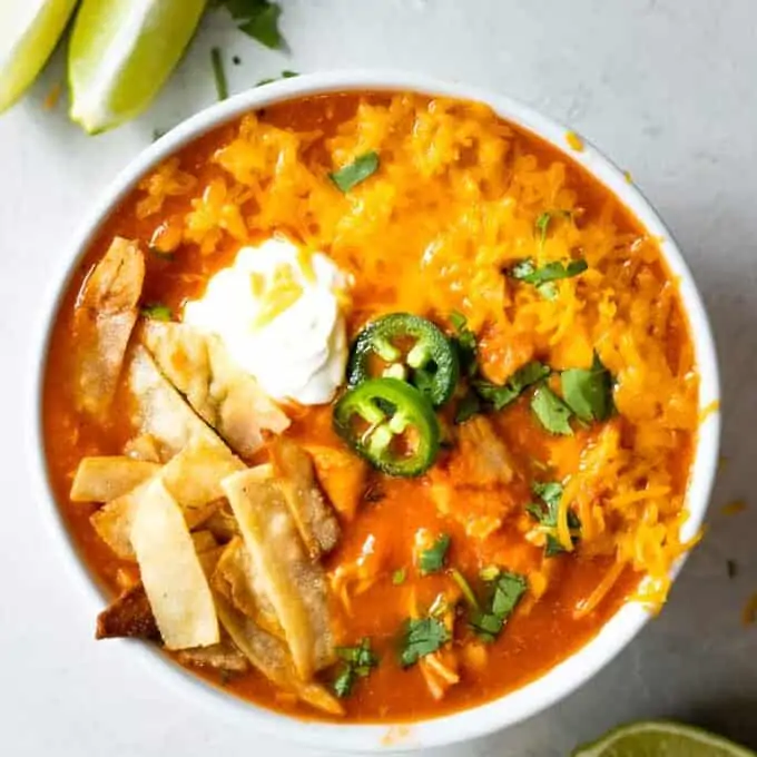 Bowl filled with chicken tortilla soup topped with melty cheese, a swirl of sour cream, sliced jalapeños and crunchy tortilla strips.