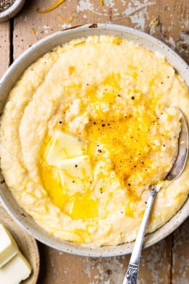 Bowl of cheese grits topped with melted butter and black pepper with a spoon.