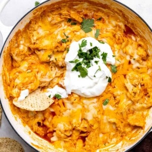 White skillet filled with cheesy chicken enchilada dip, topped with sour cream and with a tortilla chip stuck in it.