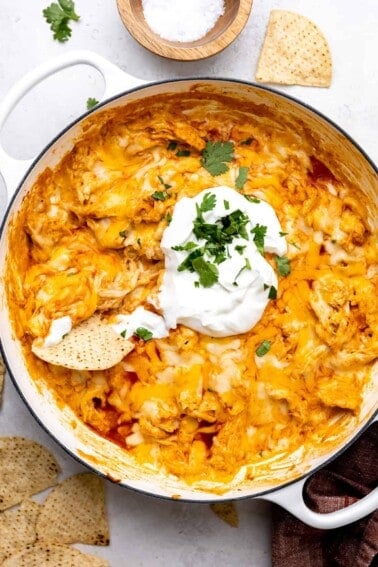 White skillet filled with cheesy chicken enchilada dip, topped with sour cream and with a tortilla chip stuck in it.