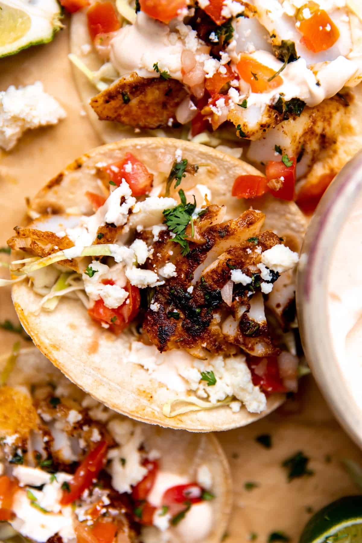 Up close of a fish taco topped with tomatoes, cabbage, crumbled cheese and fish taco sauce.