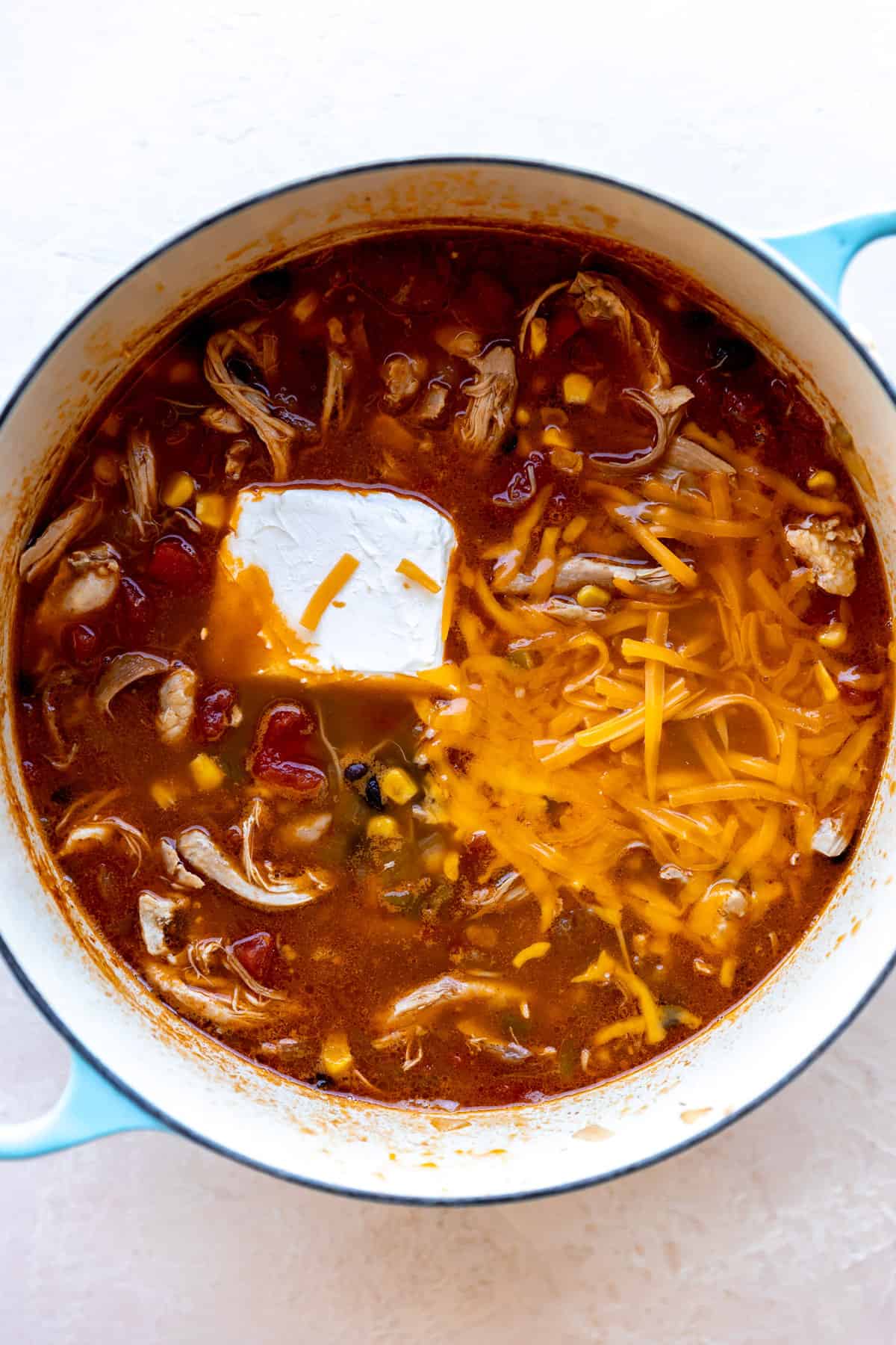 Dutch oven with chicken taco soup and and cream cheese, and shredded cheese being added to create a creamy broth.