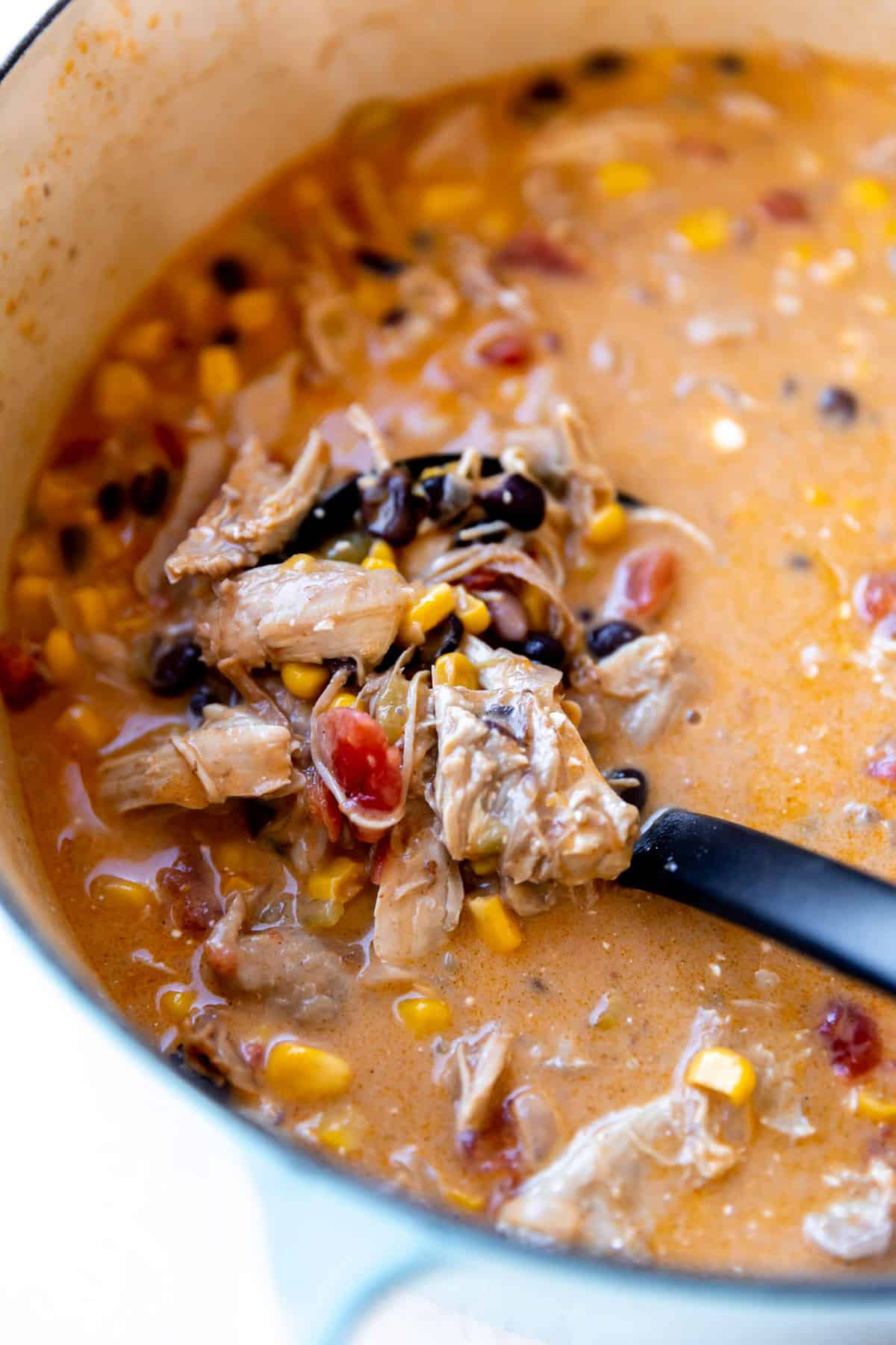 Ladle scooping up shredded chicken, beans, corn and diced tomato in a creamy broth.