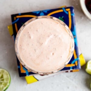 Cup filled with chipotle crema on top of a stack of mexican tiles, lime wedges around it.