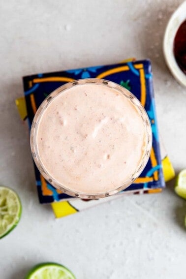 Cup filled with chipotle crema on top of a stack of mexican tiles, lime wedges around it.