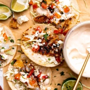 Fish tacos laying on parchment paper with lime wedges and a bowl of fish taco sauce.