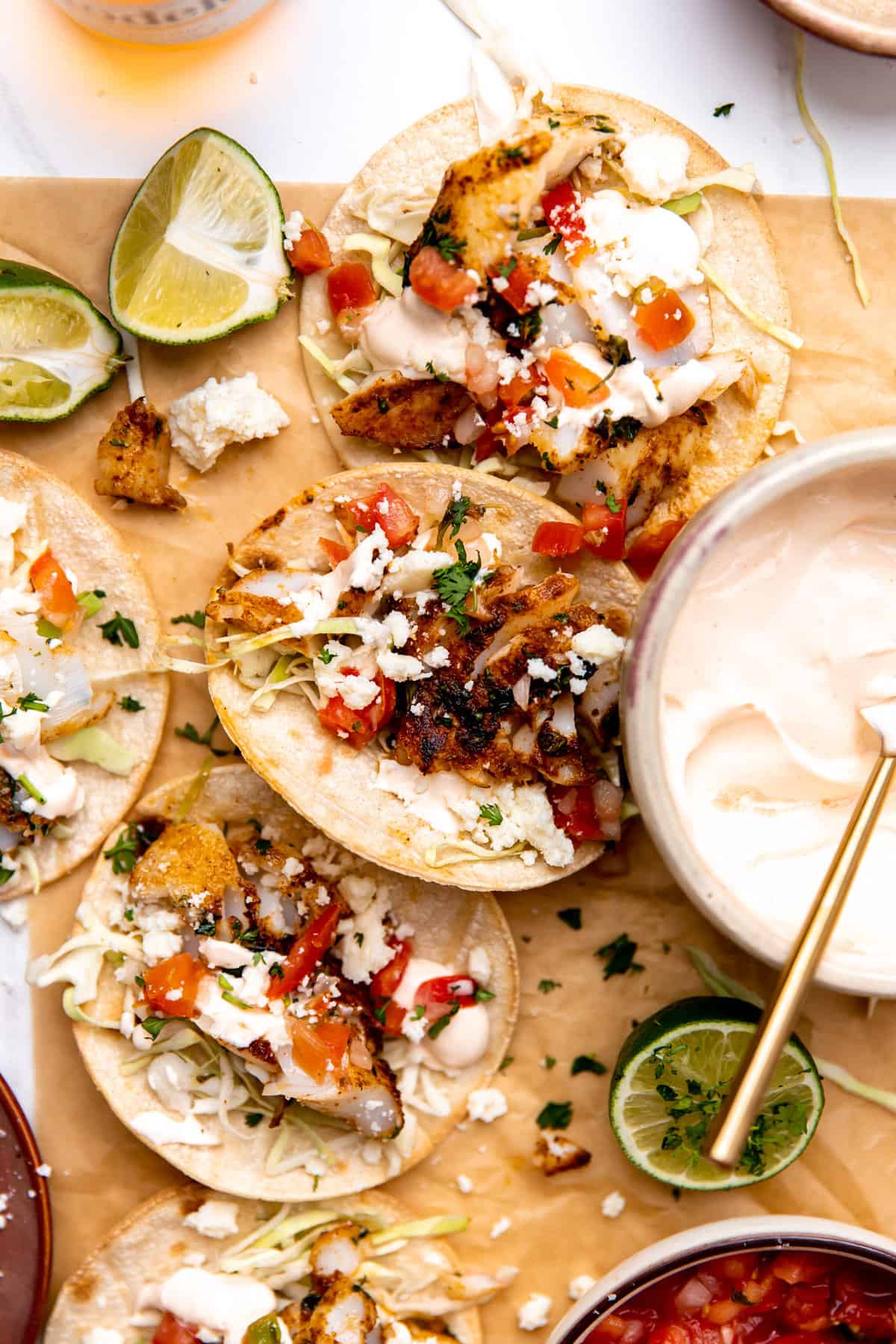 Fish tacos laying on parchment paper with lime wedges and a bowl of fish taco sauce.