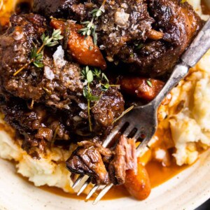 Cooked braised oxtails served over mashed potatoes, topped with thyme and cooked carrots, a fork is cutting into tender shreds of meat.