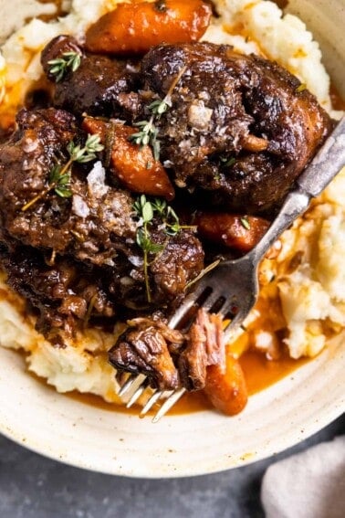 Cooked braised oxtails served over mashed potatoes, topped with thyme and cooked carrots, a fork is cutting into tender shreds of meat.