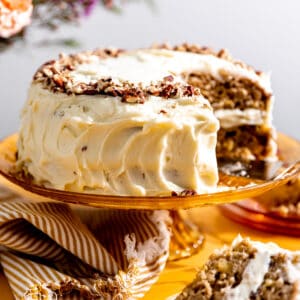 Hummingbird cake topped with pecans, on a orange glass cake stand with one slice removed and on a plate.