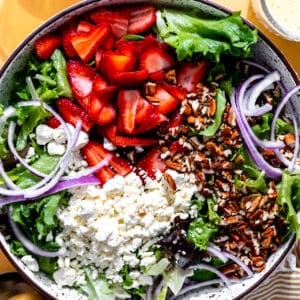 Bowl filled with salad greens topped with sliced strawberries, crumbled feta, sliced red onions and chopped pecans.