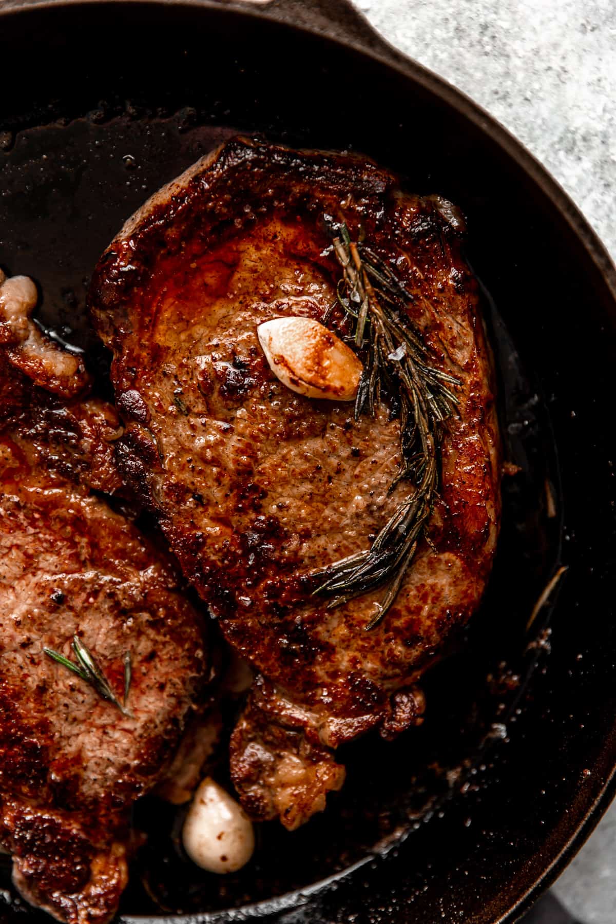 Cooked ribeye steak in a cast iron skillet topped with a sprig of rosemary and garlic cloves.