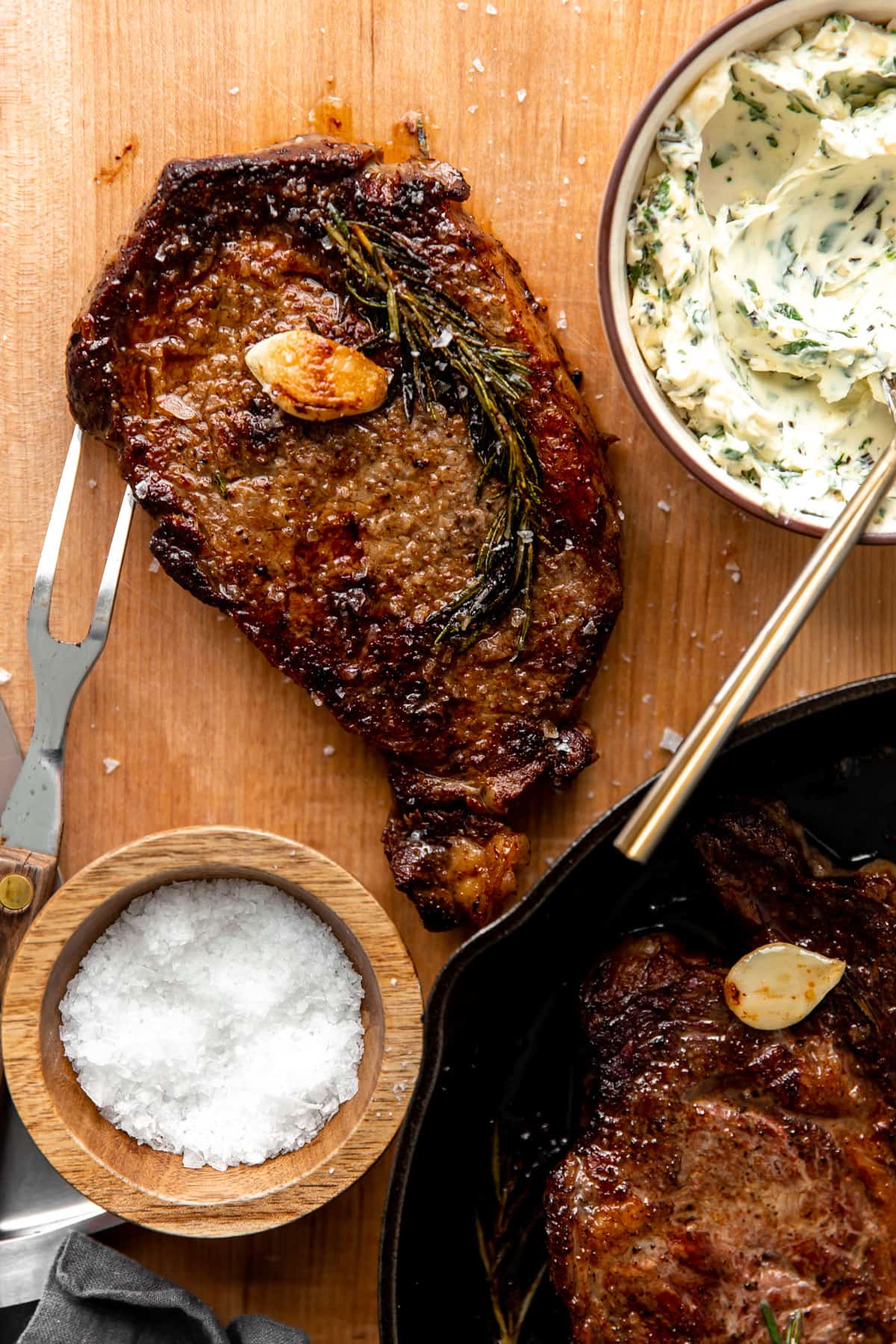 Ribeye steak after cooking resting on a cutting board with a bowl of garlic herb butter on the side. 