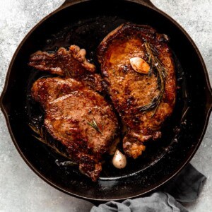 Cast iron skillet with two seared ribeyes topped with rosemary and garlic.