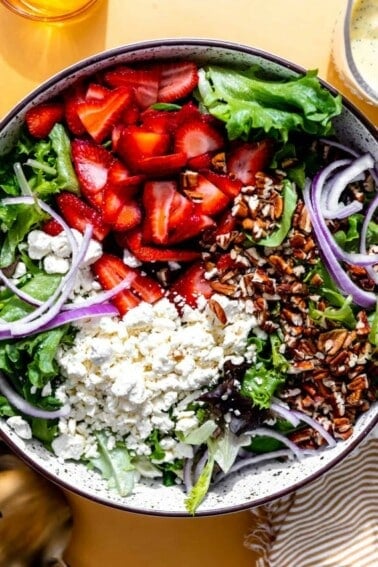 Bowl filled with salad greens topped with sliced strawberries, crumbled feta, sliced red onions and chopped pecans.