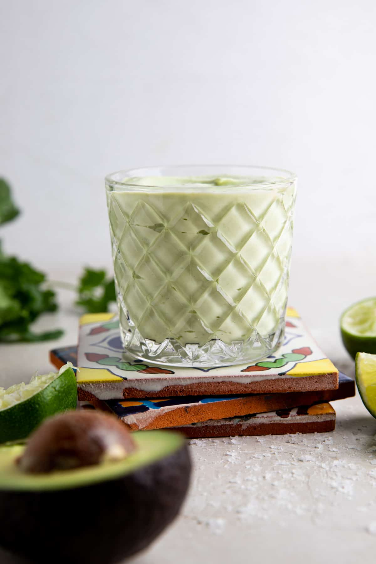 Cup with avocado crema on a stack of mexican tiles with lime wedges and avocados around it. 