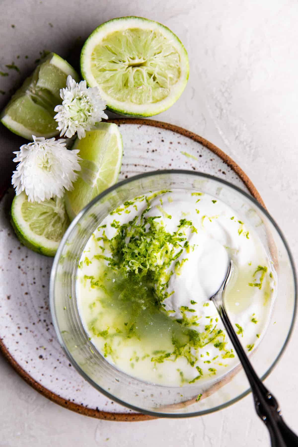 How to make Lime Crema showing a bowl filled with sour cream, lime zest and juice, minced garlic and salt. On a small plate with lime wedges and flours on the side. 