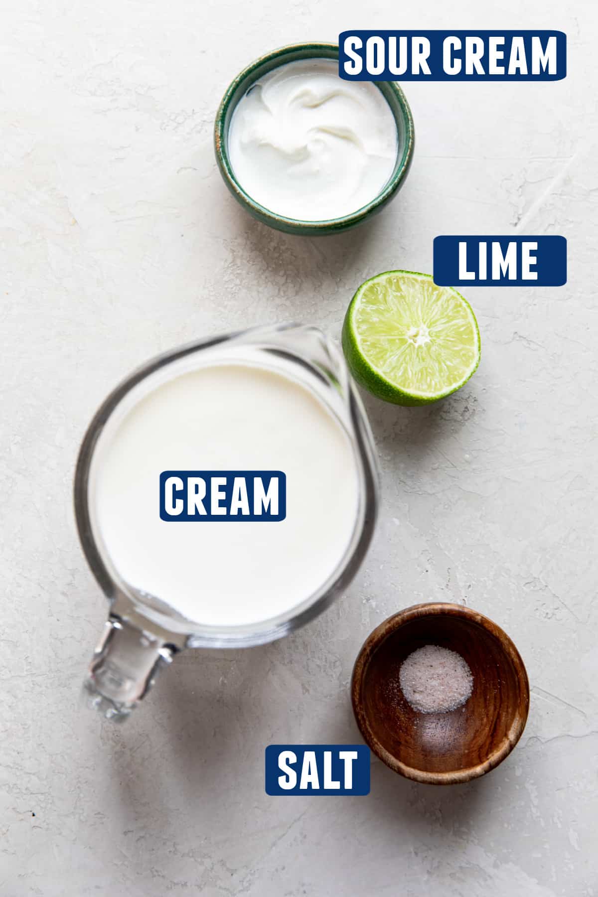 Ingredients needed to make homemade mexican crema laid out on the counter. A measuring glass of cream, bowl of sour cream, half a lime, and a small amount of salt.