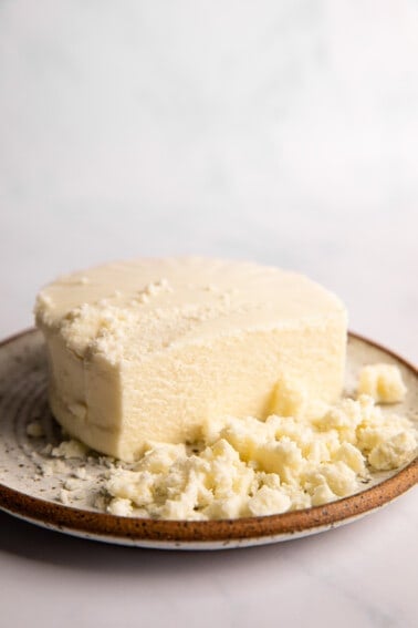 Round of queso fresco on a plate with slices cut off and crumbles.